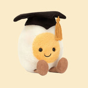 On a white background is a stuffed toy in the shape of a hard boiled egg wearing a graduation cap and smiling.