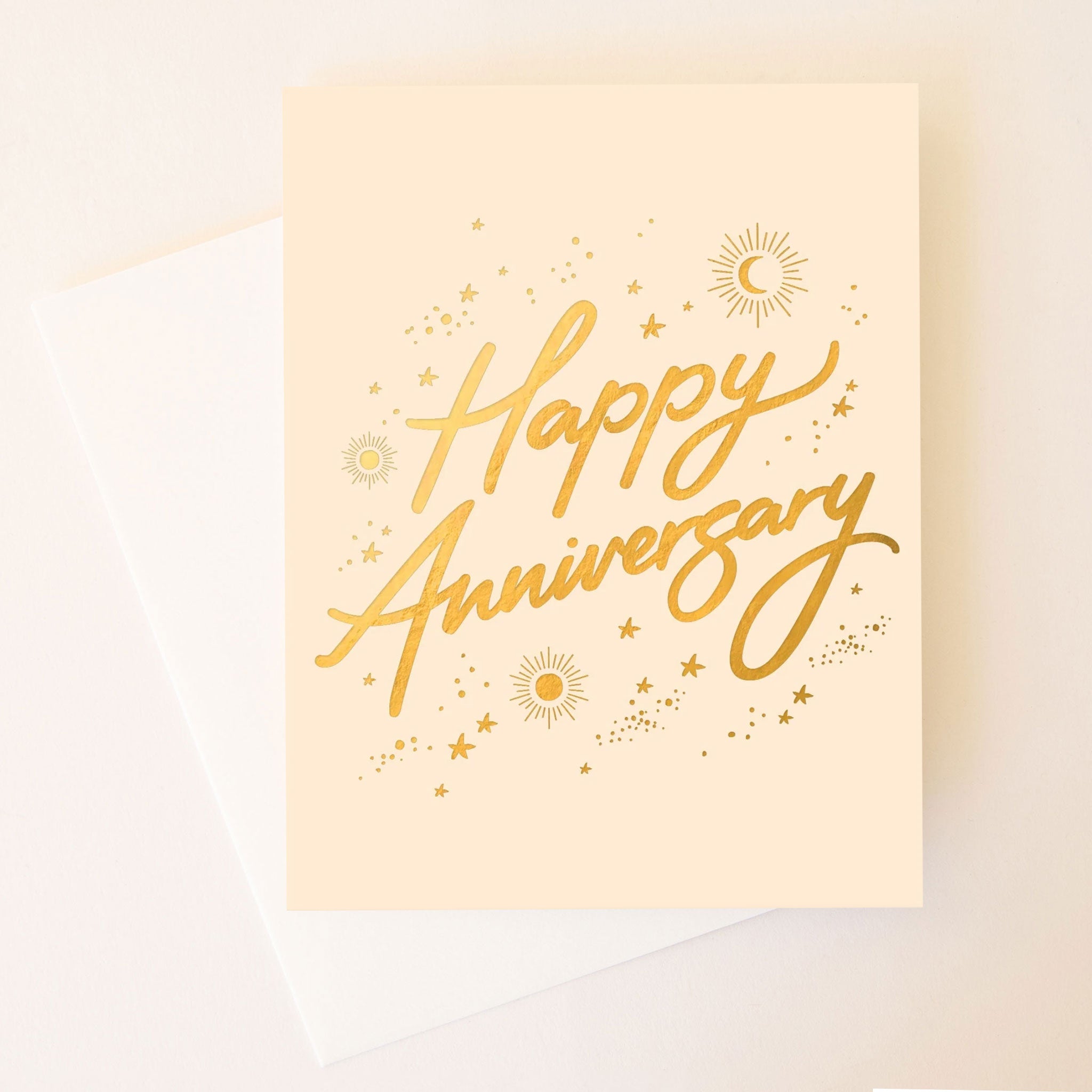 On a neutral background is a tan card with gold foil text that reads, "Happy Anniversary" along with gold foil stars, sun and moons around. 