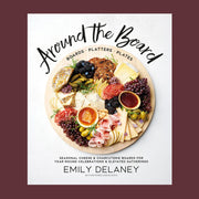 Hard cover cookbook titled 'Around the board'. The text curves around a rich, colorful cheeseboard filled with various cheeses, fruits and crackers. Within the board reads 'boards, platters, plates' in small lettering. Below the board reads 'seasonal cheese and charcuterie boards for year-round celebrations and elevated gatherings' in small black text.