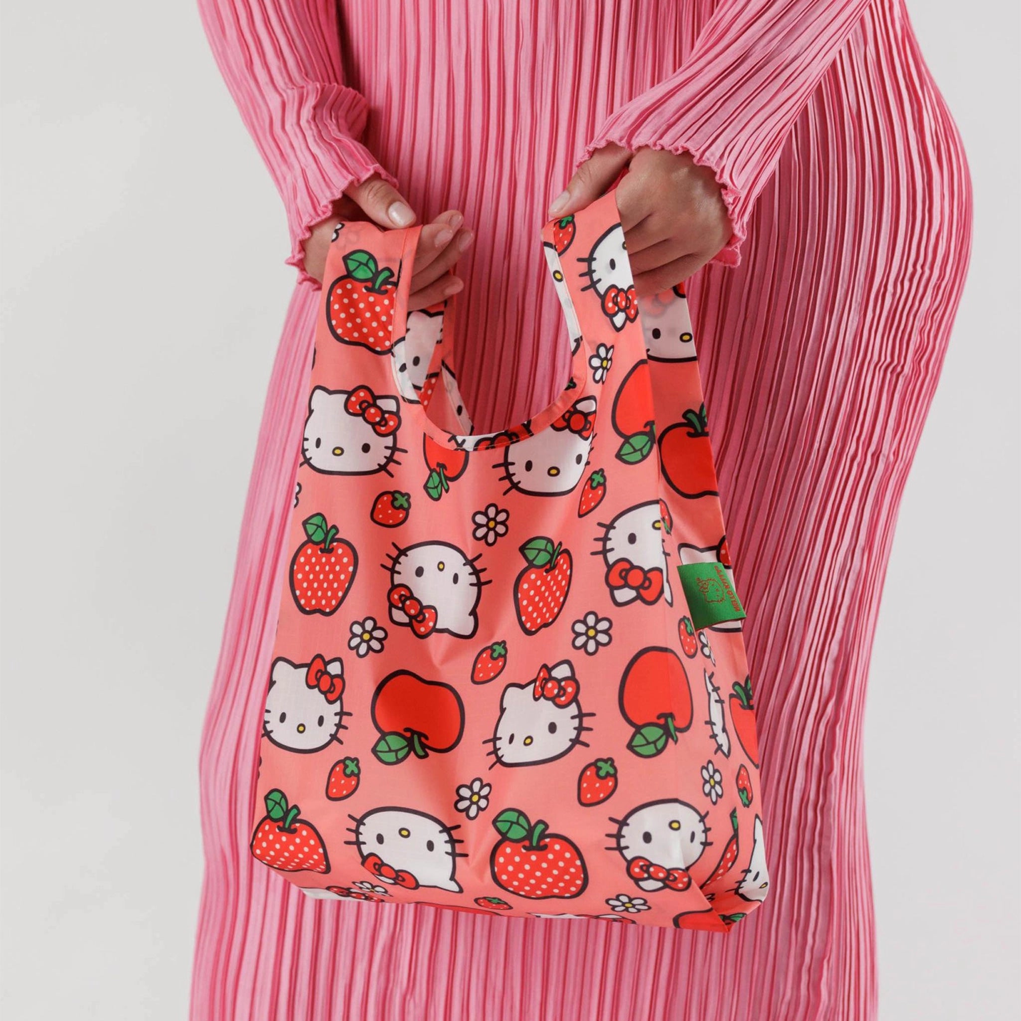 On a white background a model is holding a hello kitty and apple printed nylon tote bag.