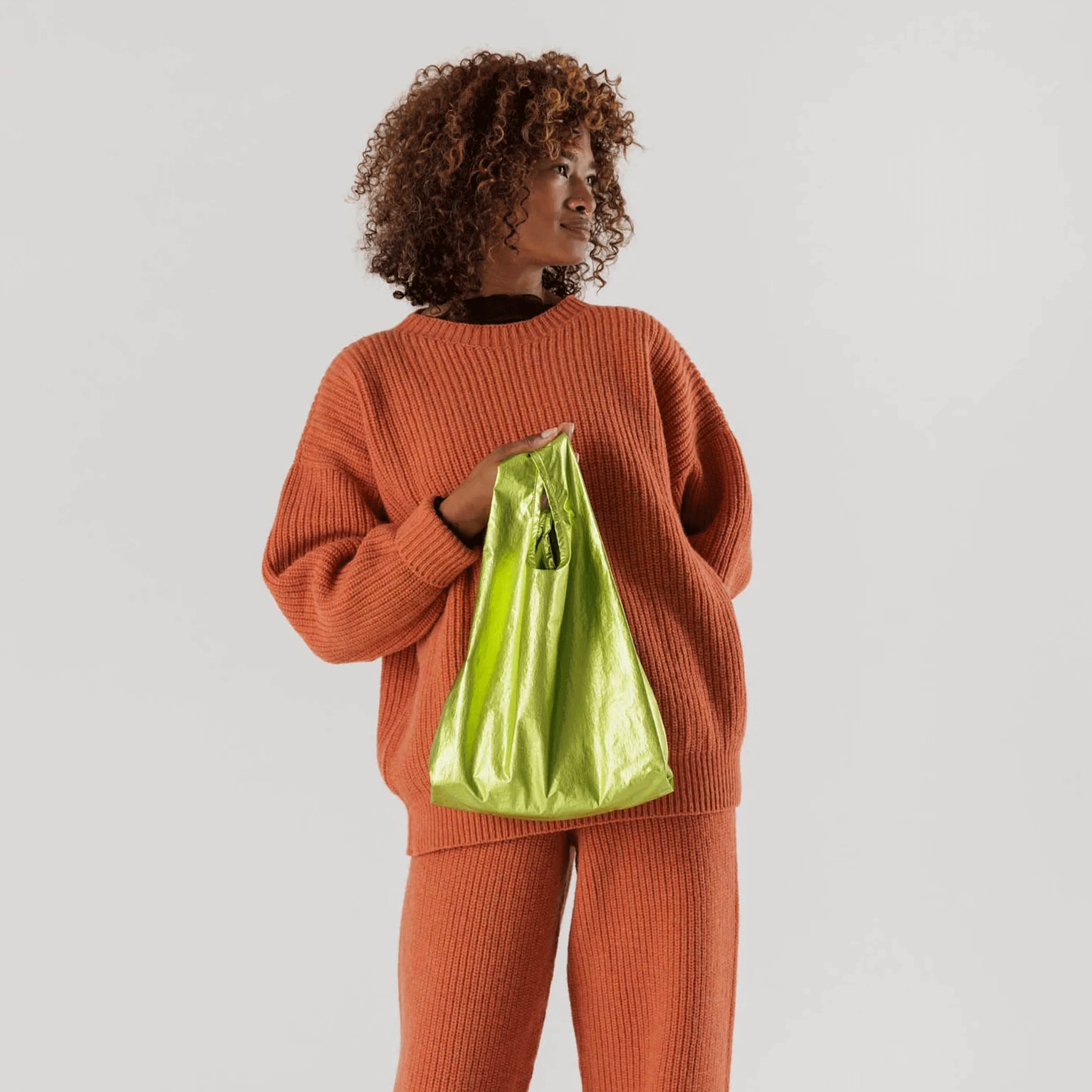 On a cream background is a model holding a neon green metallic nylon bag. 