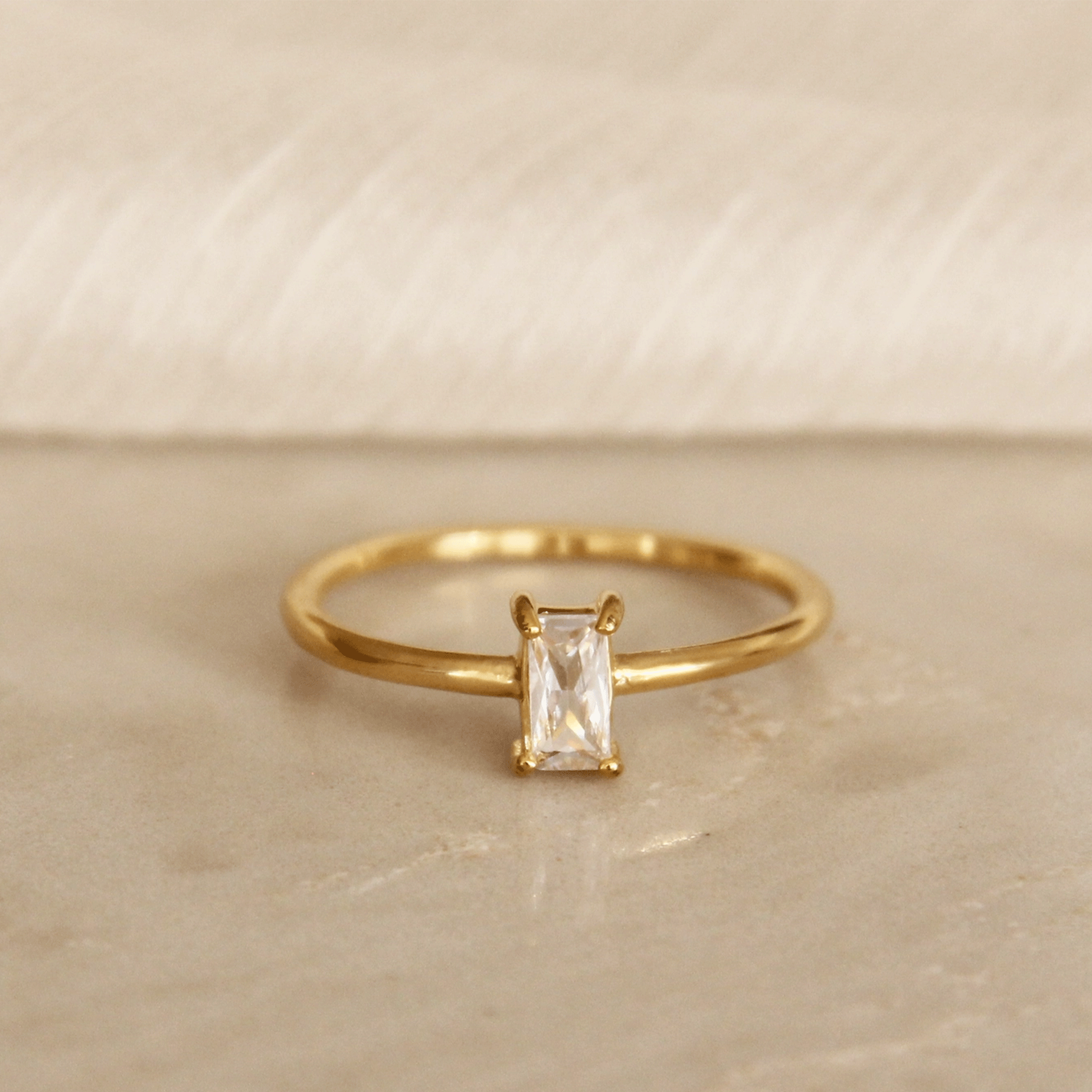 On a tan background is a thin gold band ring with a solitaire baguette shaped cz stone in the center. 