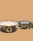 Round tin candle vessel with dark and light gold floral pattern and white wax candle with three wicks. Candle vessel lid has text, "Voluspa. Baltic Amber."