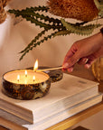 Round tin candle vessel with dark and light gold floral pattern and white wax candle with three wicks. Candle vessel lid has text, "Voluspa. Baltic Amber."