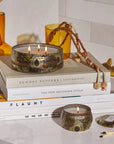 Round tin candle vessel with dark and light gold floral pattern and white wax candle with three wicks. Candle vessel lid has text, "Voluspa. Baltic Amber."