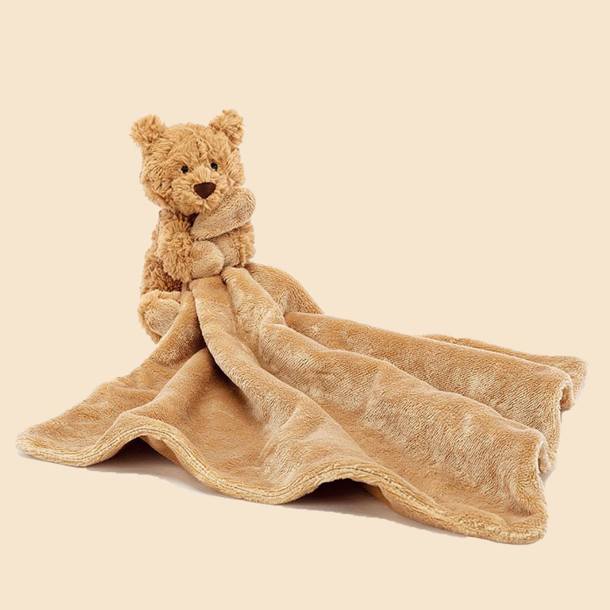 On a tan background is a tan cuddly stuffed toy bear with a small square blanket attached that can roll up for on the go. 