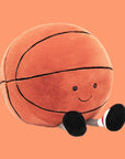 On an orange background is an orange basketball stuffed toy with a smiling face and feet.