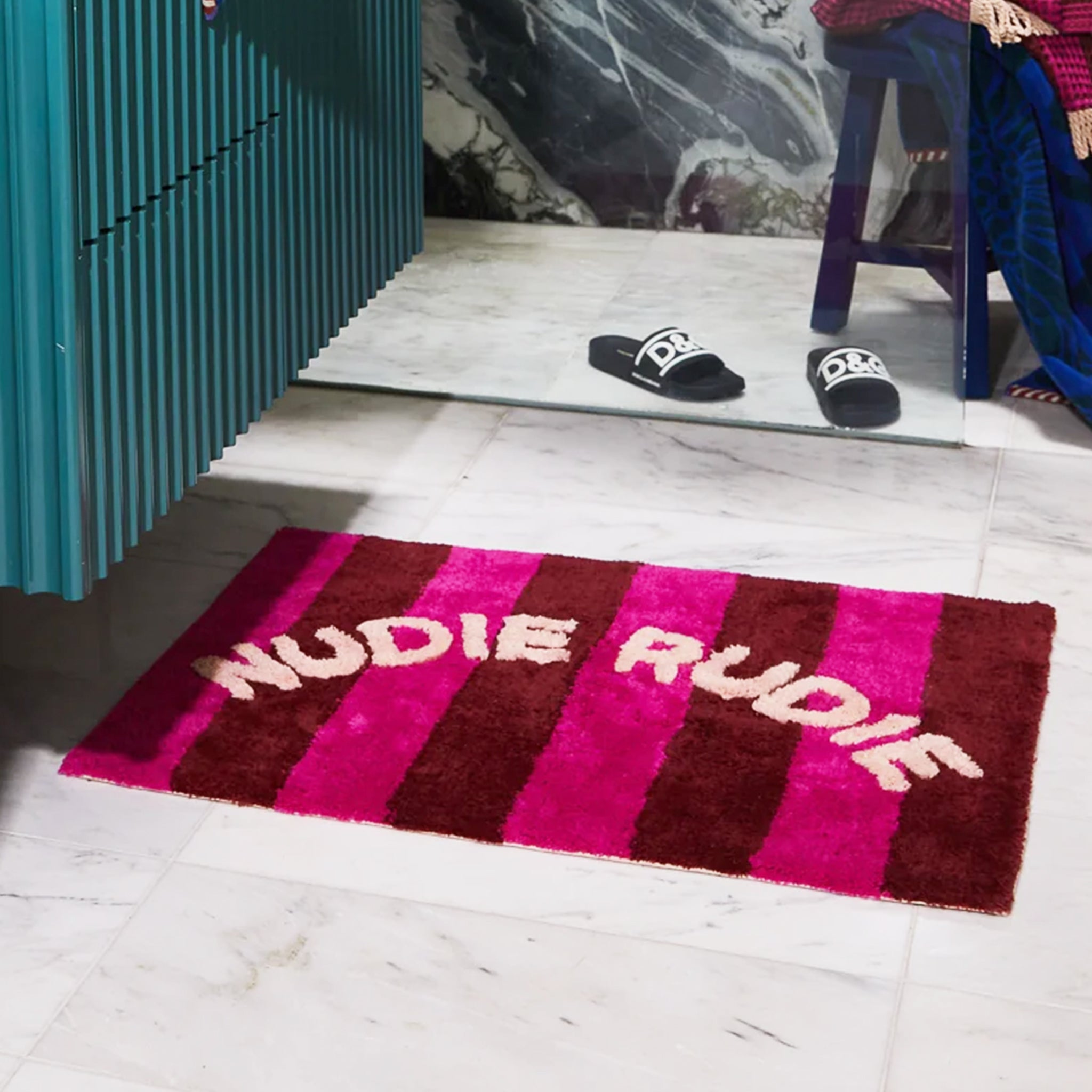 A hot pink and burgundy striped bath mat with white text arched in the center that reads, &quot;Nudie Rudie&quot;.