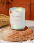 An orange and vibrant pink iridescent glass jar filled with a candle along with a white square label on the front that repeats the notes of the candle as well as the name, "Beach Vacay" in gold letters.
