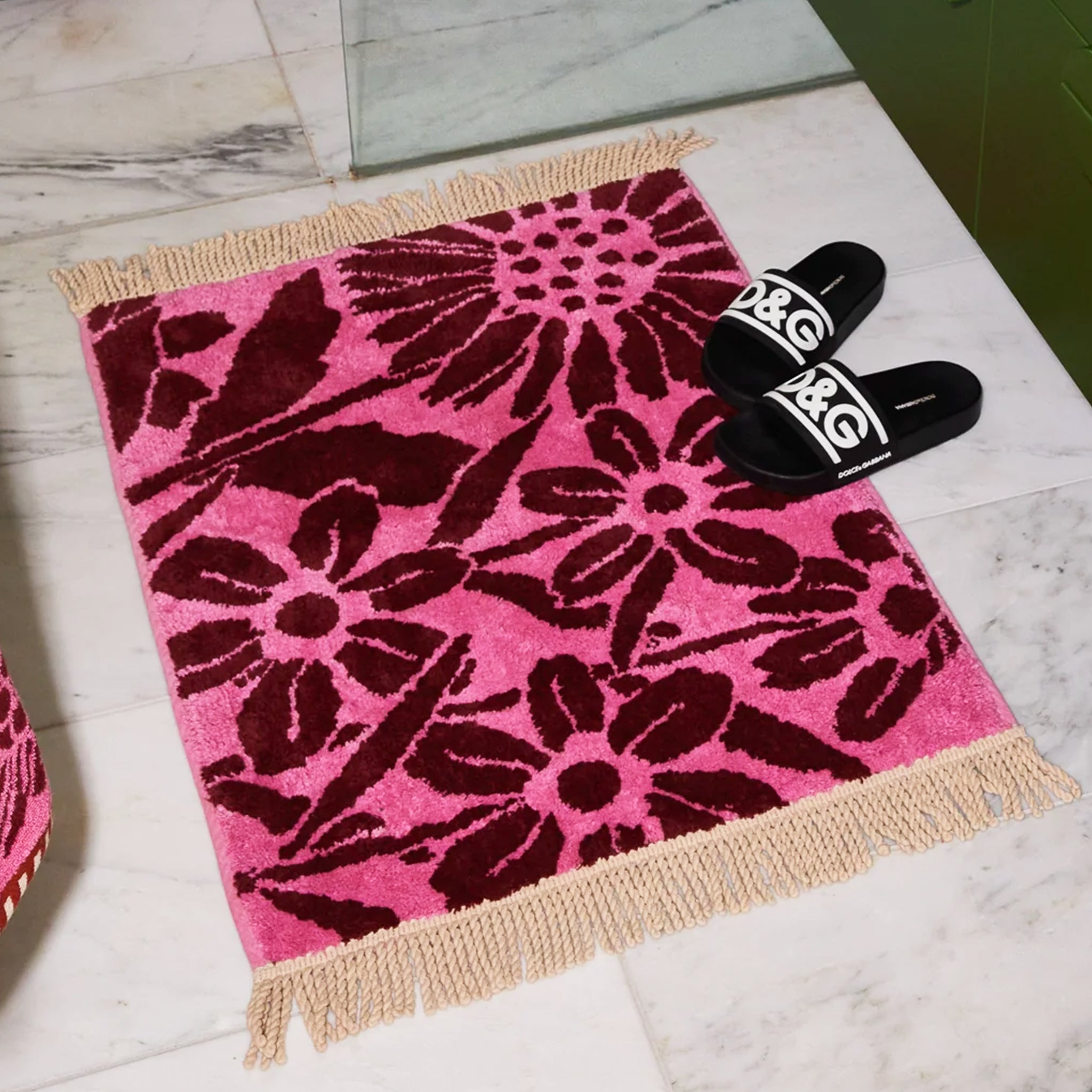 A pink and dark burgundy floral print bath mat with twist fringe details on two ends. 