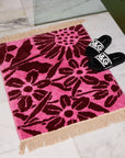 A pink and dark burgundy floral print bath mat with twist fringe details on two ends.