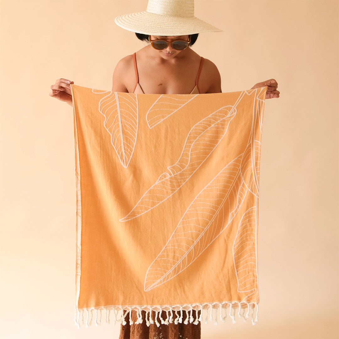 On a tan background is an orange beach towel with a banana leaf print and white tassels on each end. 