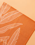 On an orange background is an orange kitchen towel with a white banana leaf design.