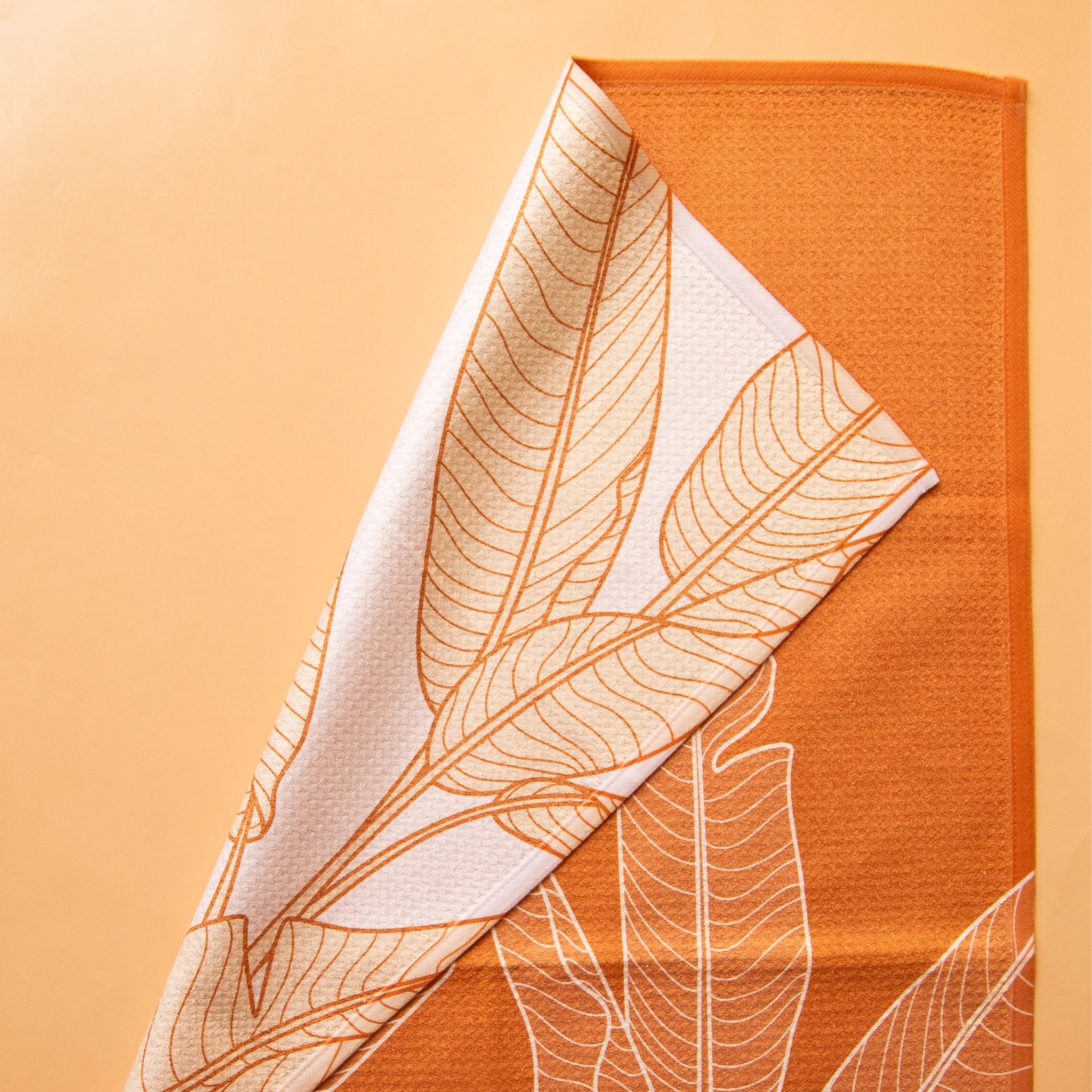 On an orange background is an orange kitchen towel with a white banana leaf design.