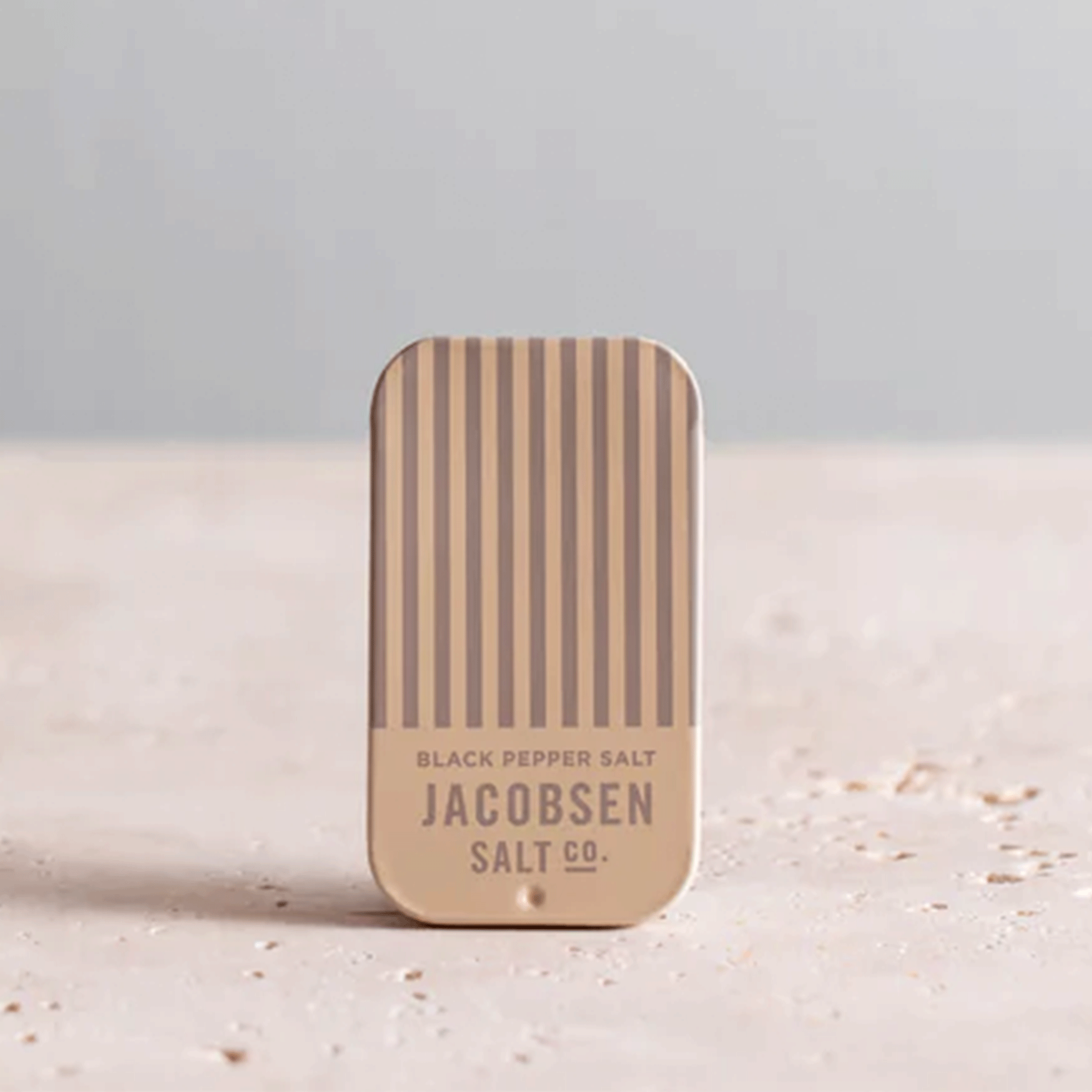 A tan and grey striped tin that reads, &quot;Black Pepper Salt Jacobsen Salt Co.&quot;.