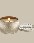On a cream background is a light silver and gold mini tin candle with white wax and a single wick. Also included is the matching lid.