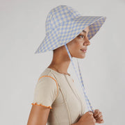 On a white background is a flexible sun hat with straps for securing around the neck with a blue and white gingham print. 