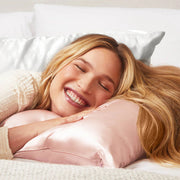 A smiling woman with long blonde hair rests her head on a blush colored satin pillow.