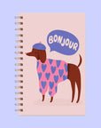 A light pink journal with an illustration of a dog wearing a beret and a speech bubble above that reads, "Bonjour".