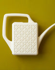An ivory plastic watering can with a narrow spout and square handle and a rectangle breeze block design on the sides.