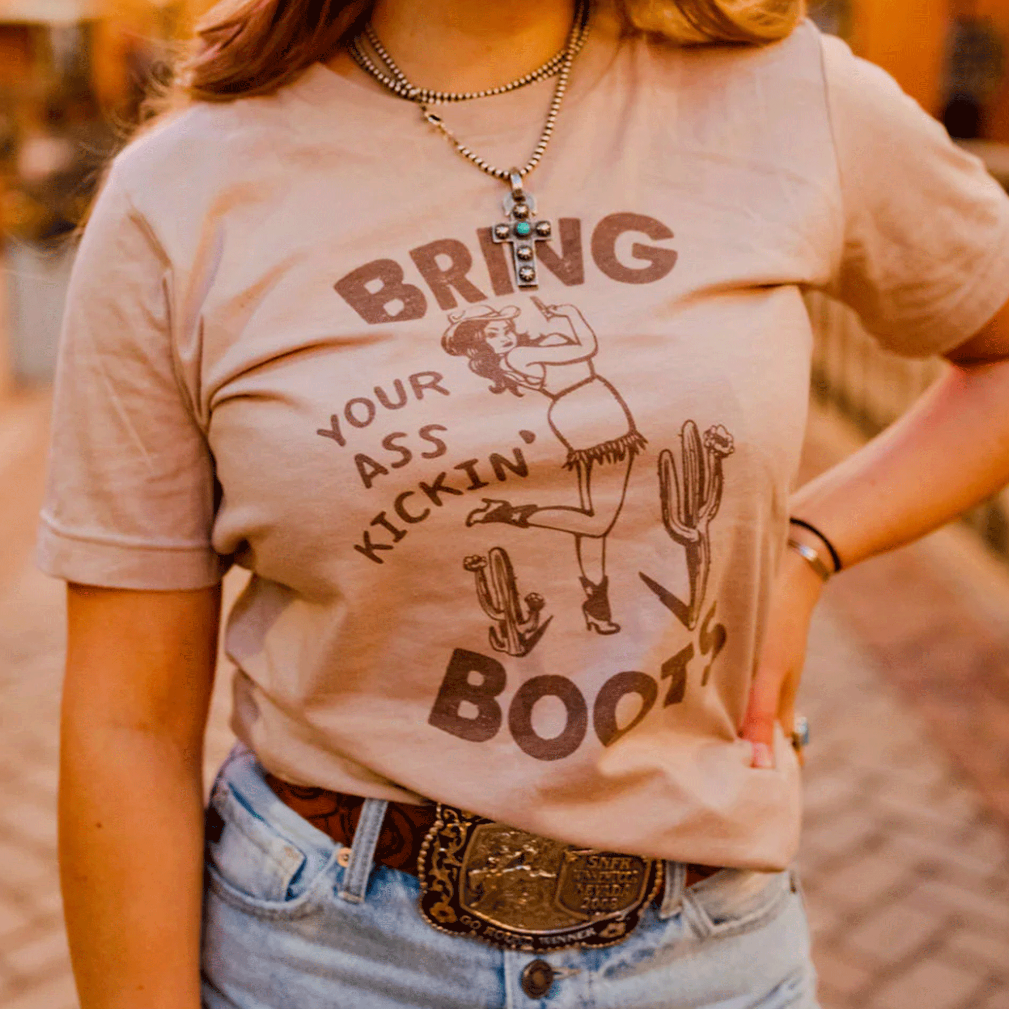 A model wearing a tan graphic tee with a design featuring a woman dressed in western style clothing in the middle of two cactus and text around that reads, &quot;Bring Your Ass Kickin&#39; Boots&quot;.