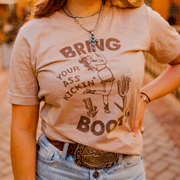A model wearing a tan graphic tee with a design featuring a woman dressed in western style clothing in the middle of two cactus and text around that reads, "Bring Your Ass Kickin' Boots".