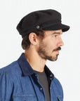 On a white background is a model wearing a black fiddler cap with a cord detail.