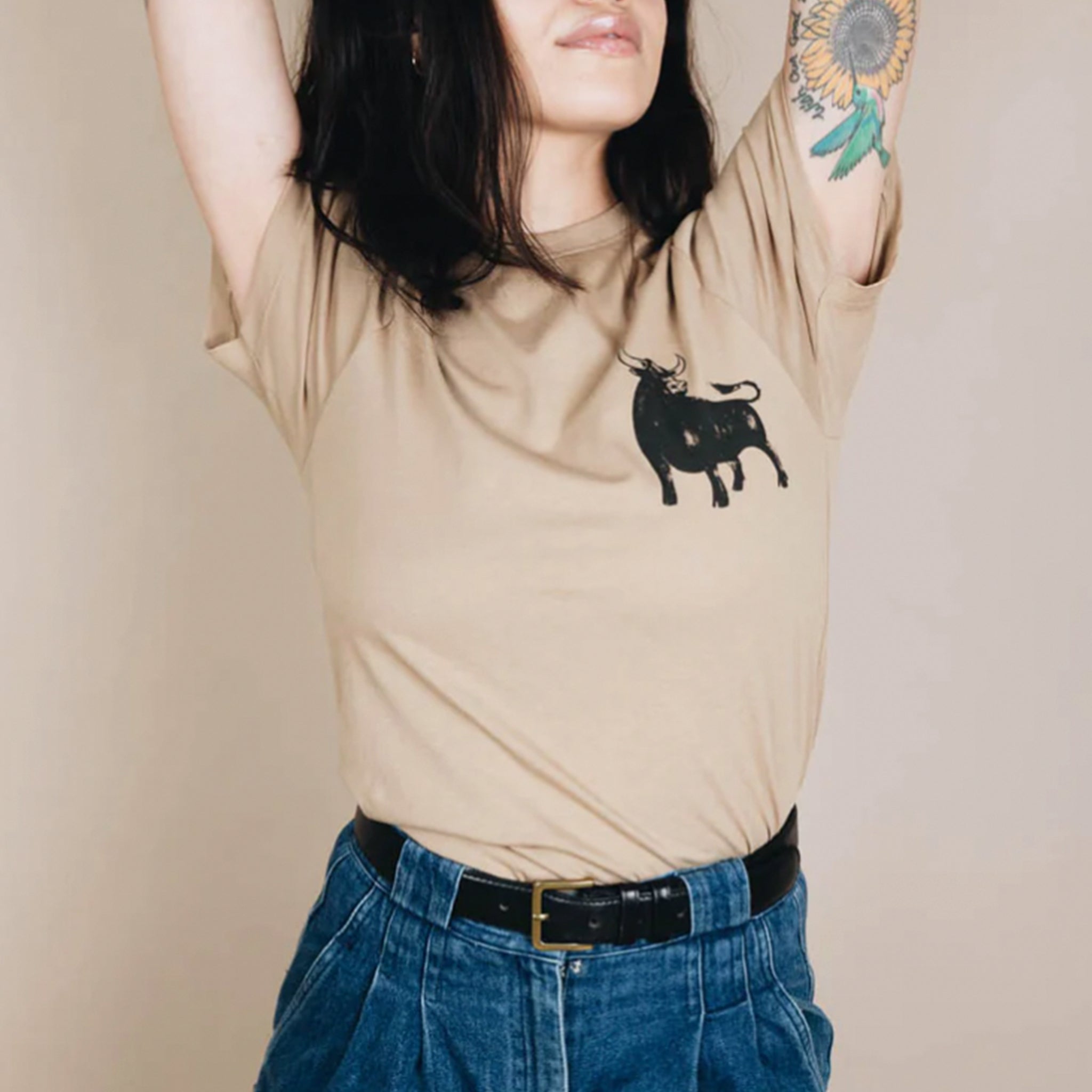 On a tan background is a tan t-shirt with a black bull screen printed on the front and text on the back that reads, &quot;Bull Shirt&quot;. 