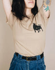 On a tan background is a tan t-shirt with a black bull screen printed on the front and text on the back that reads, "Bull Shirt".