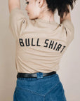 On a tan background is a tan t-shirt with a black bull screen printed on the front and text on the back that reads, "Bull Shirt".