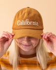 On a peachy background is a model wearing a toffee colored corduroy snapback hat that has white text embroidered across the front reading, "California".