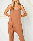 On a white background is a model wearing a camel colored jumpsuit with a drop crotch, front pockets and a v-neck.