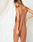 On a white background is a model wearing a camel colored jumpsuit with a drop crotch, front pockets and a v-neck.