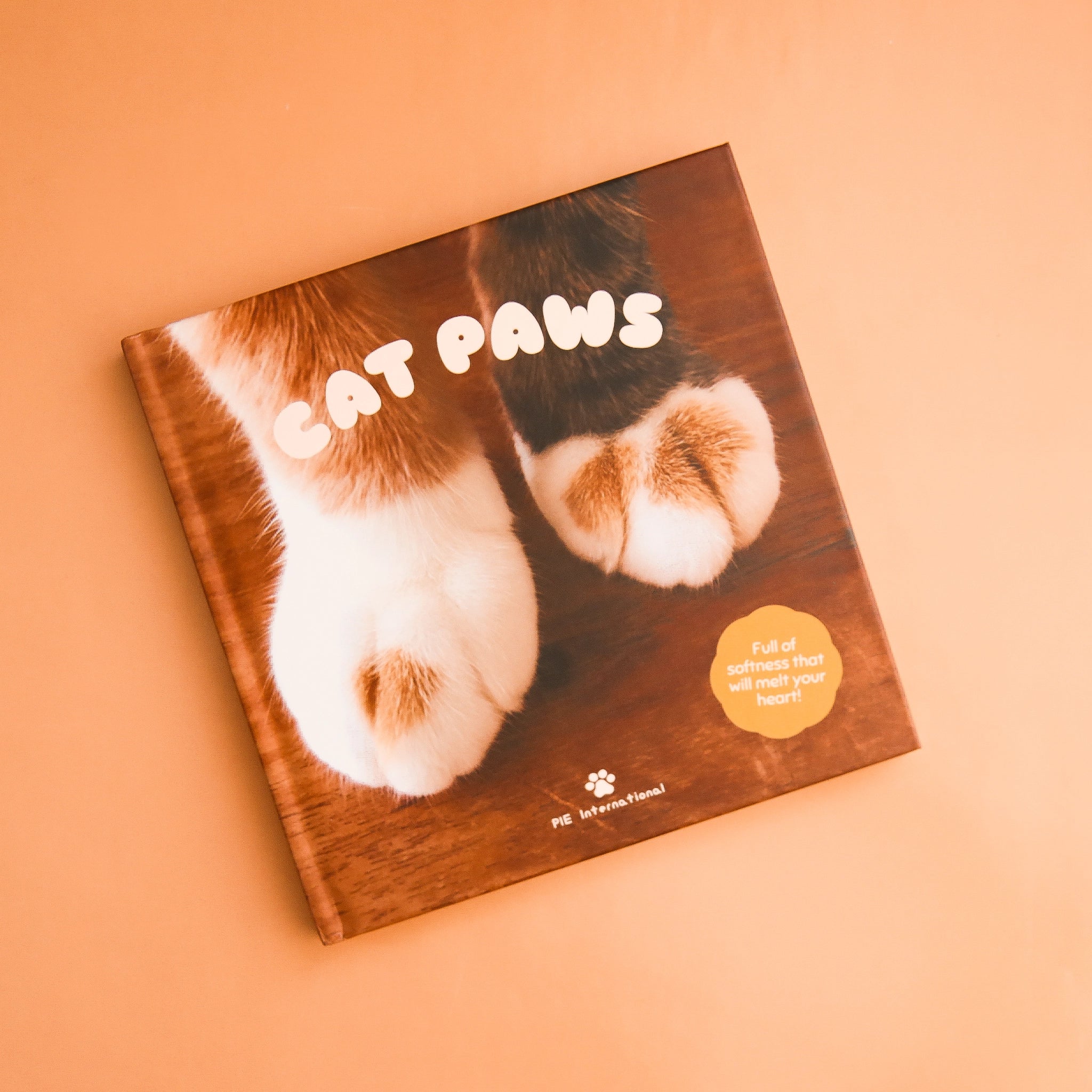 On a peach background is a book cover featuring a photograph of cat paws against a wood floor along with the title, "Cat Paws", "Full of softness that will melt your heart!".