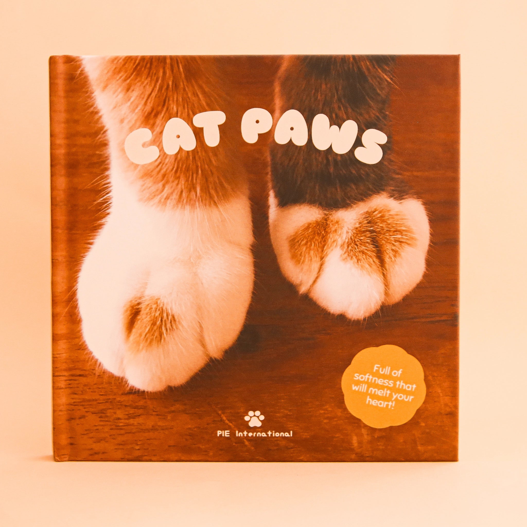 On a peach background is a book cover featuring a photograph of cat paws against a wood floor along with the title, &quot;Cat Paws&quot;, &quot;Full of softness that will melt your heart!&quot;.