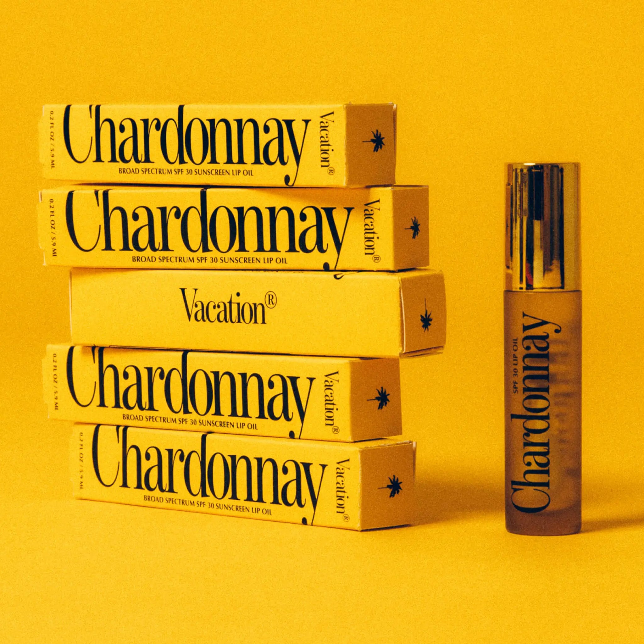 A clear bottle of the Chardonnay lip oil featuring a doe foot applicator next to stacked boxes of the lip oil in the packaging that reads, "Chardonnay Broad Spectrum SPF 30 Sunscreen Lip Oil".