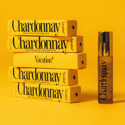 A clear bottle of the Chardonnay lip oil featuring a doe foot applicator next to stacked boxes of the lip oil in the packaging that reads, "Chardonnay Broad Spectrum SPF 30 Sunscreen Lip Oil".