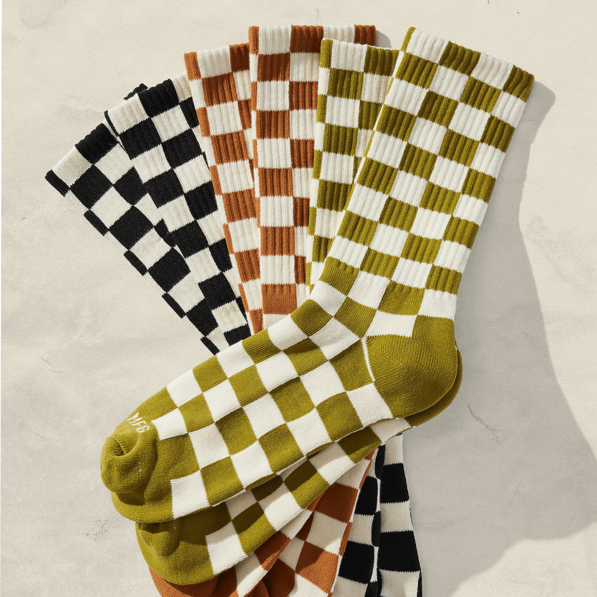 On a cream background is a pair of green and ivory checkered socks alongside a rust pair and a black and white. 
