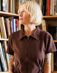 A model standing in front of a bookcase with a chocolate brown utility jumpsuit on with a zipper going up the front, a collar and a cinched waist.