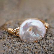 A white pearl ring with gold wire band sitting in sand.