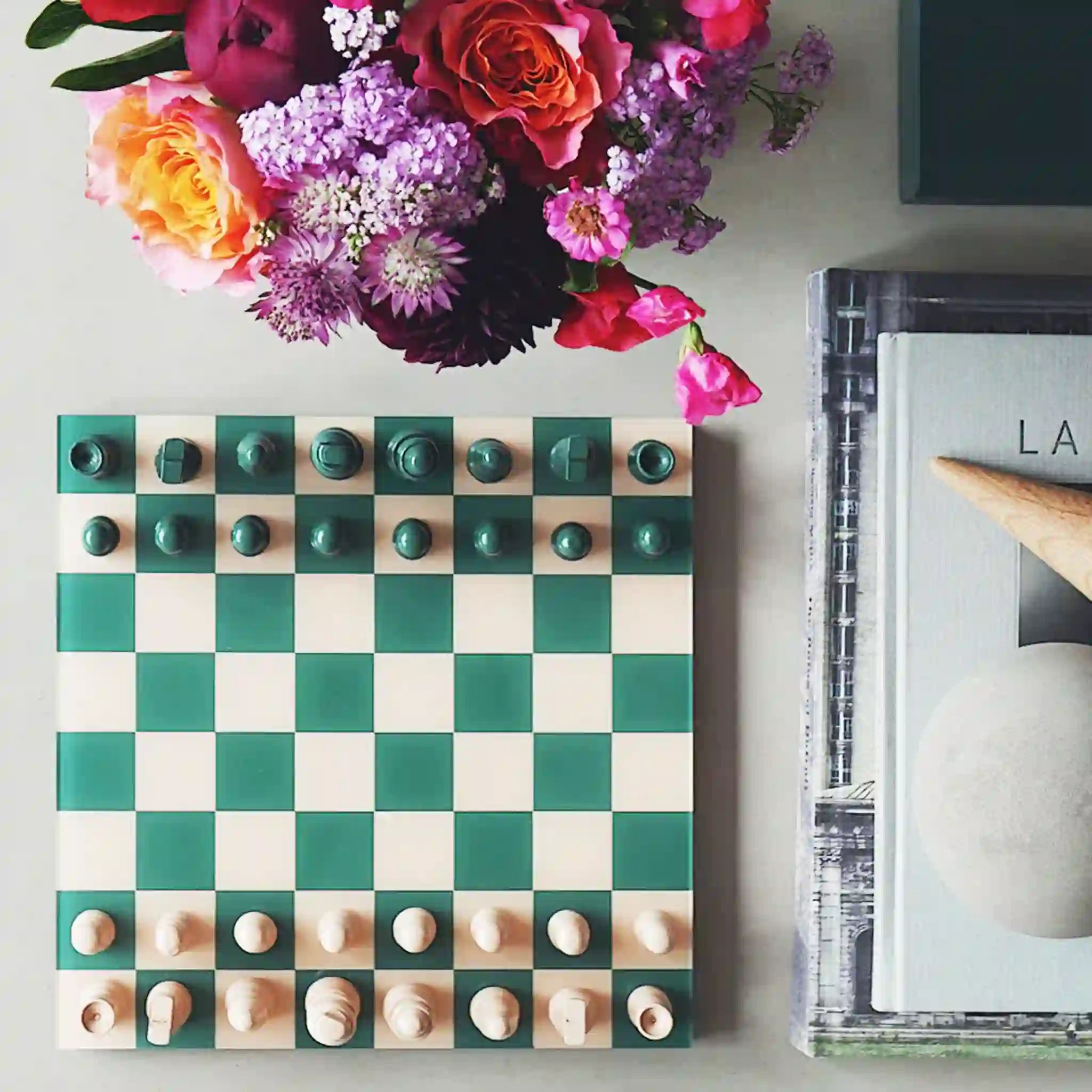 A green and cream colored chess board set staged on a coffee table with flowers and books. 