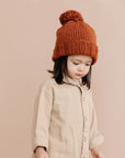 On a peachy background is a model wearing a reddish colored knit beanie with a pom pom on top.