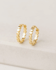 On a tan background is a pair of gold hoop earrings with a dainty circle detailing on the hoops.