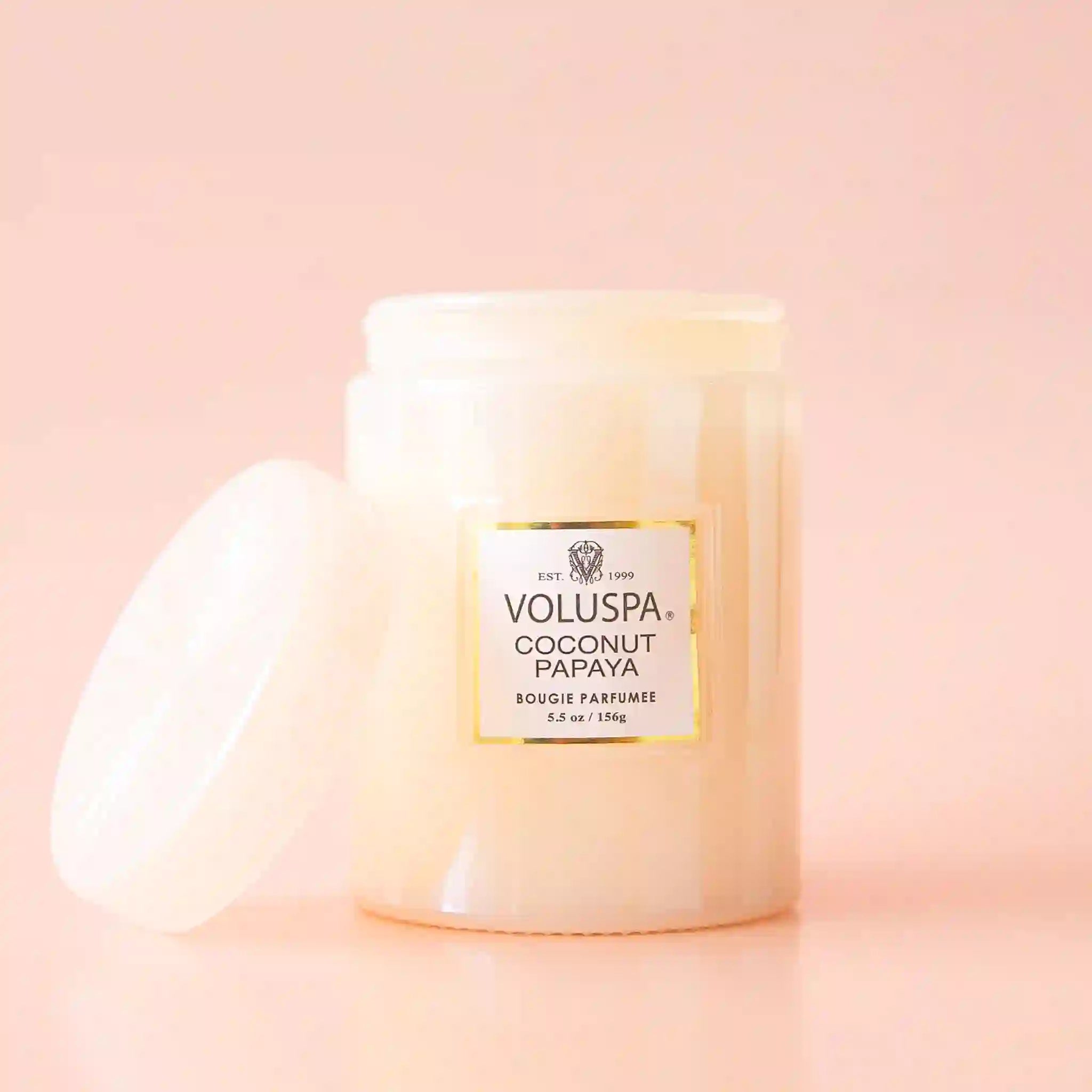 On a neutral background is a light pink glass jar candle with a white label with &quot;Voluspa Coconut Papaya&quot;. 