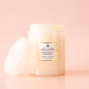 On a neutral background is a light pink glass jar candle with a white label with "Voluspa Coconut Papaya". 