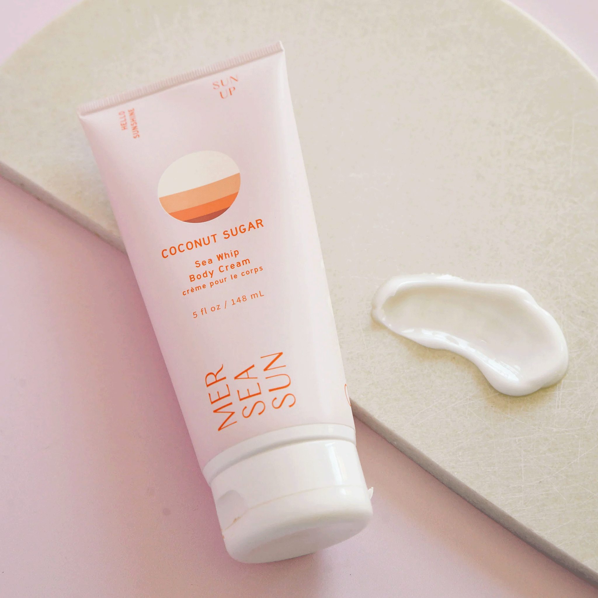 On a pink and white background is a light pink bottle of lotion with orange text on the front that reads, &quot;Mersea Sun Coconut Sugar Sea Whip Body Cream&quot;. 