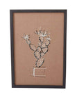 On a white background is a brown piece of wall art with white and black stitching of a cactus in a pot. The art comes in a simple black frame.