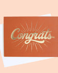 Deep orange colored greeting card with a gold foil starburst design surrounding the word congrats.