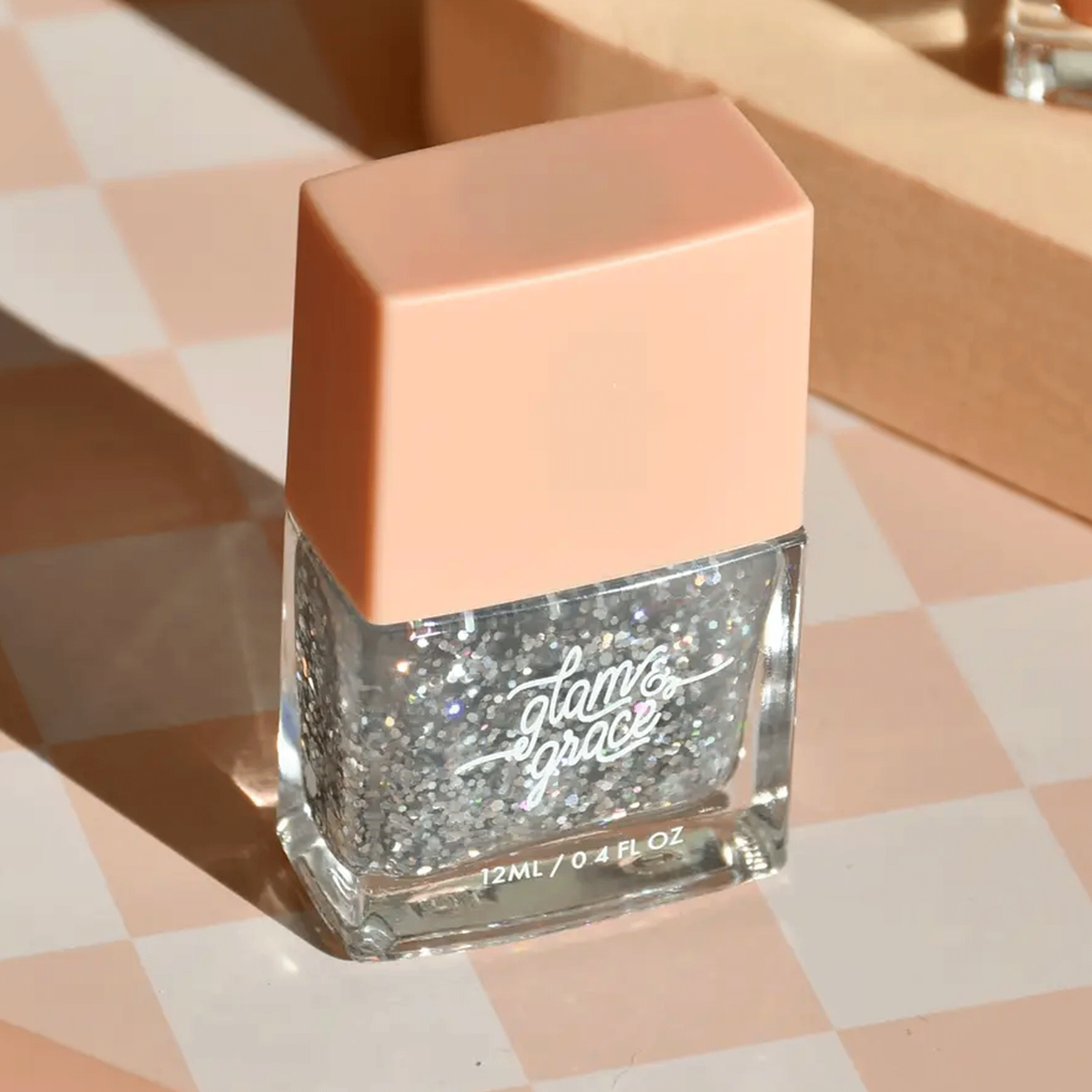 On a ivory and peach checkered surface is a rectangle bottle of silver sparkle nail polish with white text that reads, &quot;glam &amp; grace&quot; on the front. 