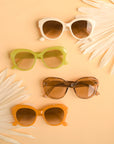 On a peachy background is a group photo of the different color options that the Donna Sunglasses come in. There is a white pair, a green, a light brown and an orange.